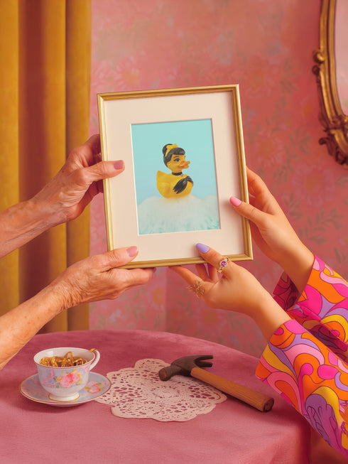 A chic rubber duck styled like Audrey Hepburn’s Holly Golightly, wearing a high bun, yellow bow, black gloves, and a sparkly choker. The duck floats in a pile of foam and is framed in white on a bubbly bathroom shelf with a red tile backdrop and soap dispenser.